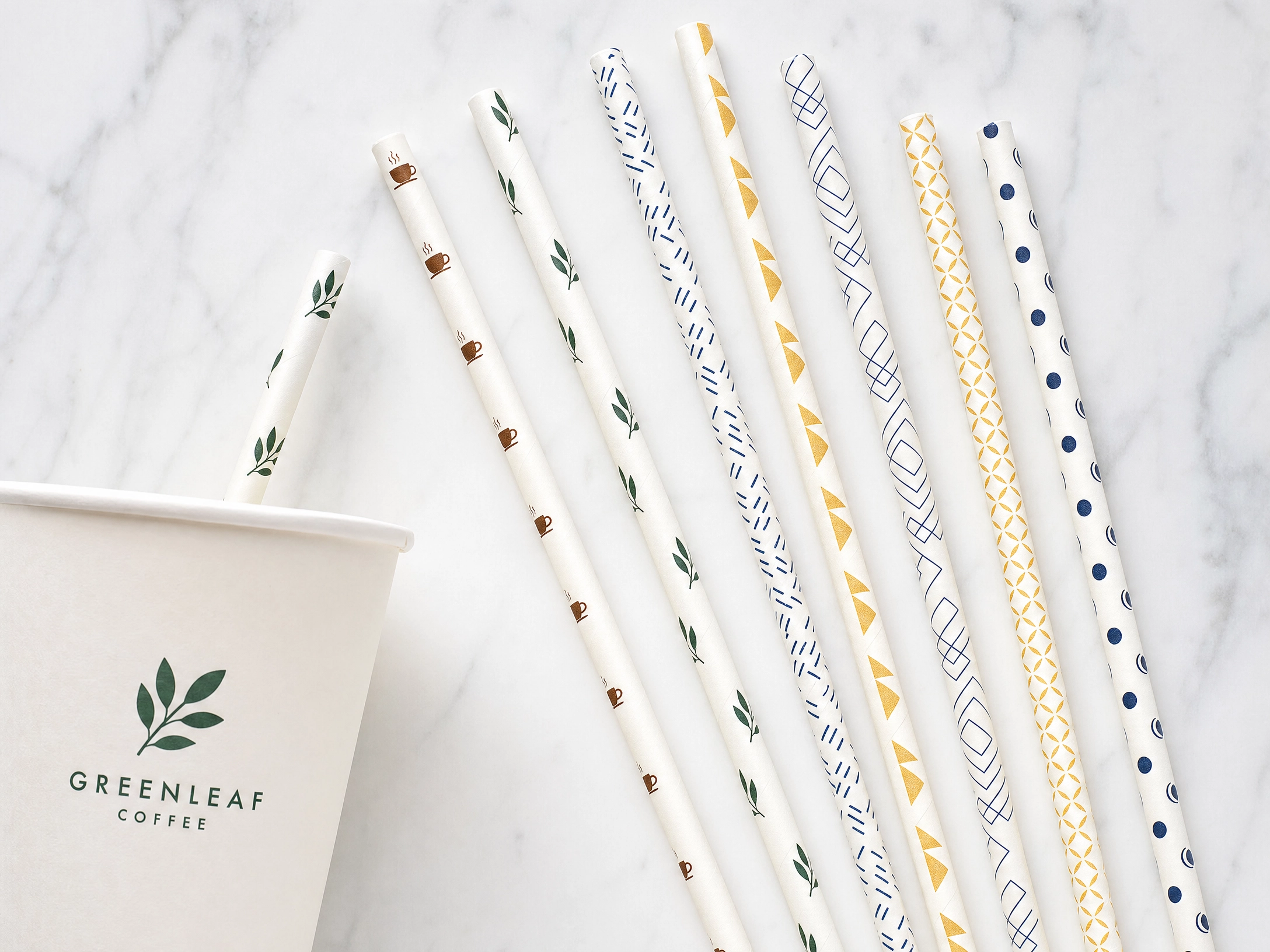 Custom printed paper straws with brand logo patterns on white background, fanned out beside a branded coffee cup on marble — custom printed straws wholesale